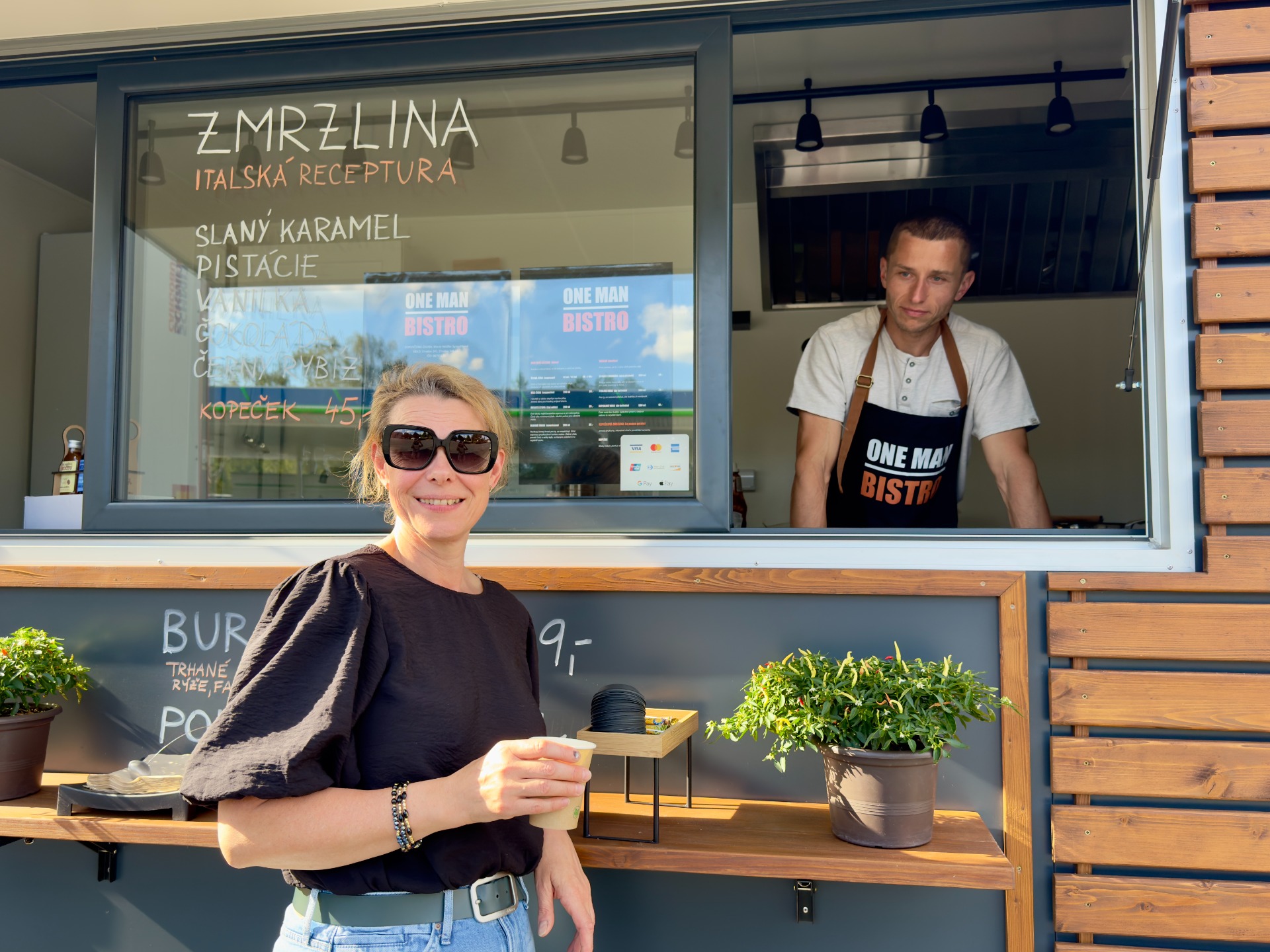 Usměvavá žena v popředí před okénkem bistro, za sklem muž v zástěře One Man Bistro.