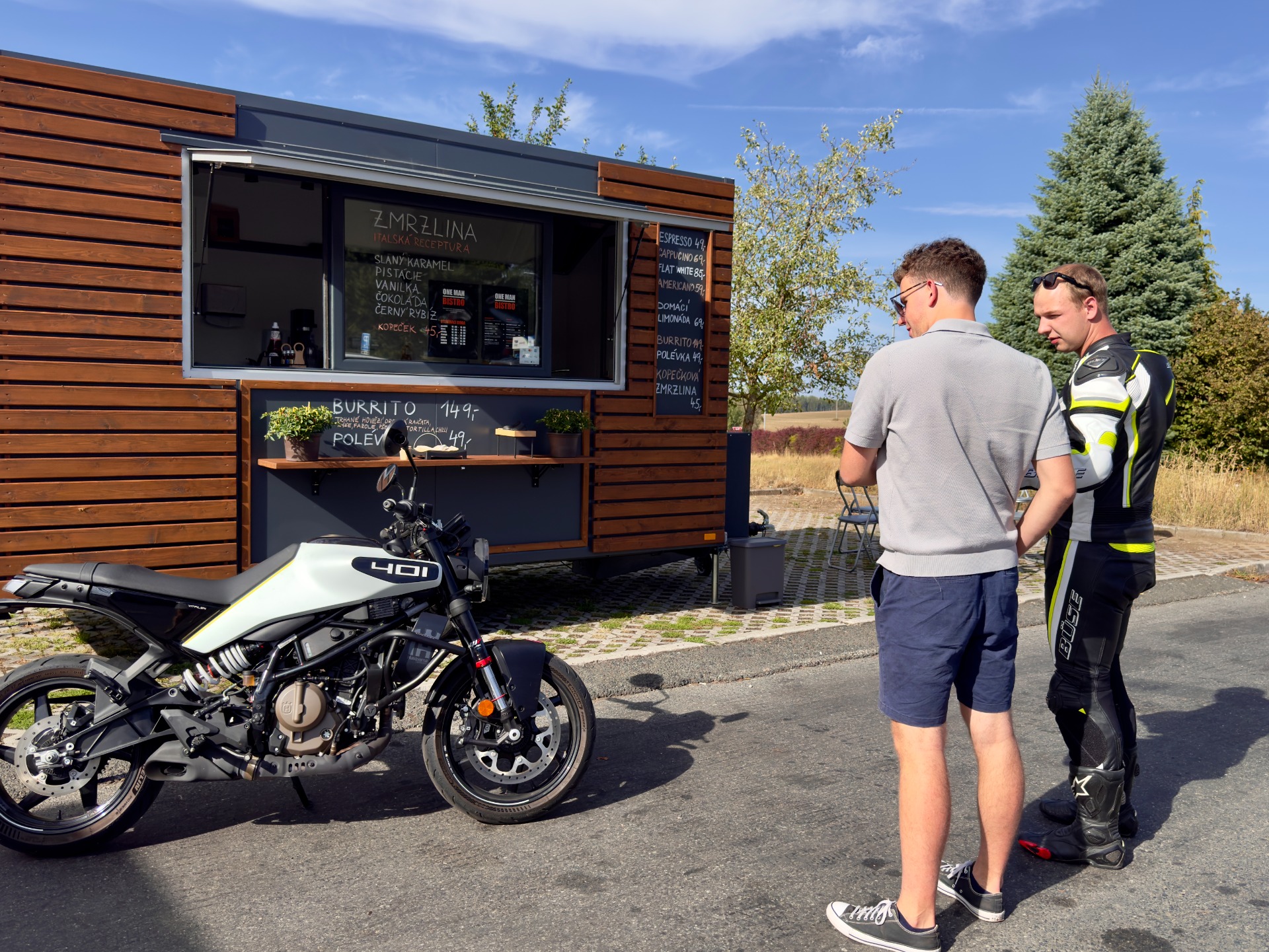 Dva muži stojí u pojízdného bistra s nápisy, vedle motocyklu na slunečném parkovišti.